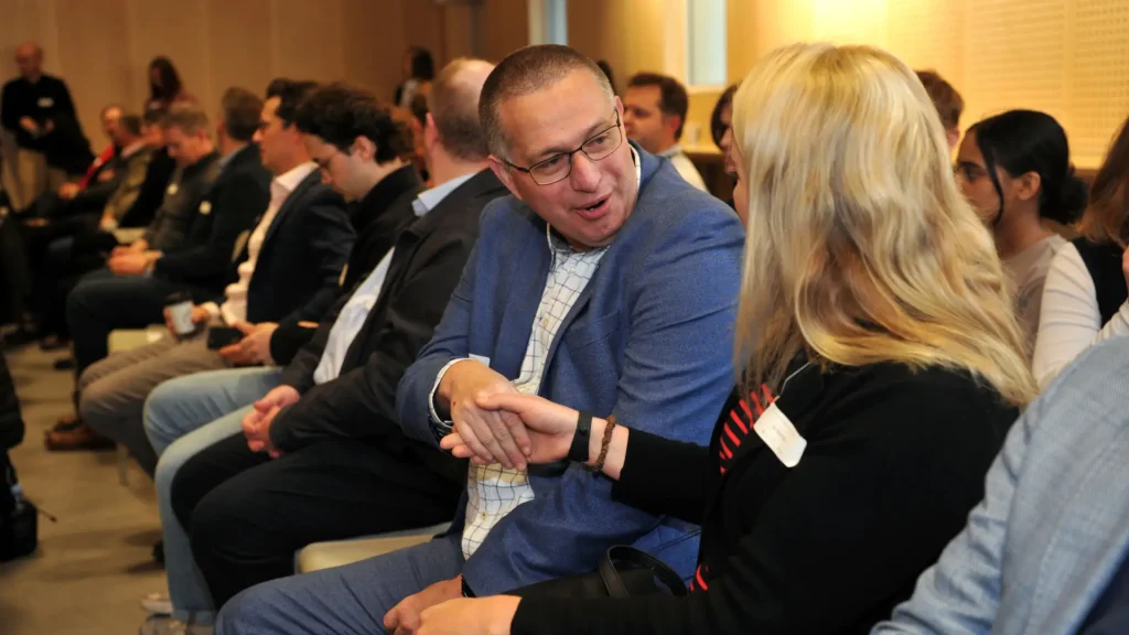 A man in a blue blazer enthusiastically shaking hands and conversing with a woman seated next to him.
